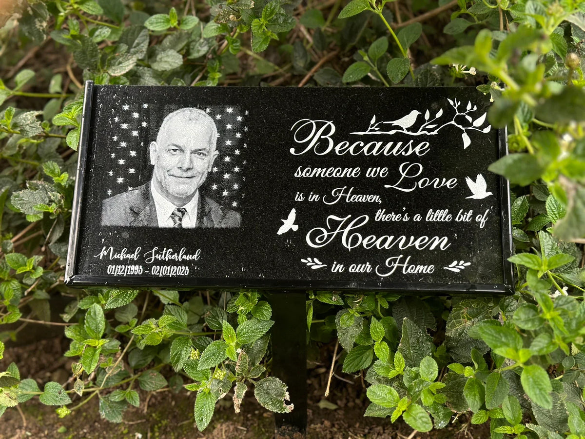 Personalized black granite memorial plaque with a portrait and white inscription displayed among garden plants