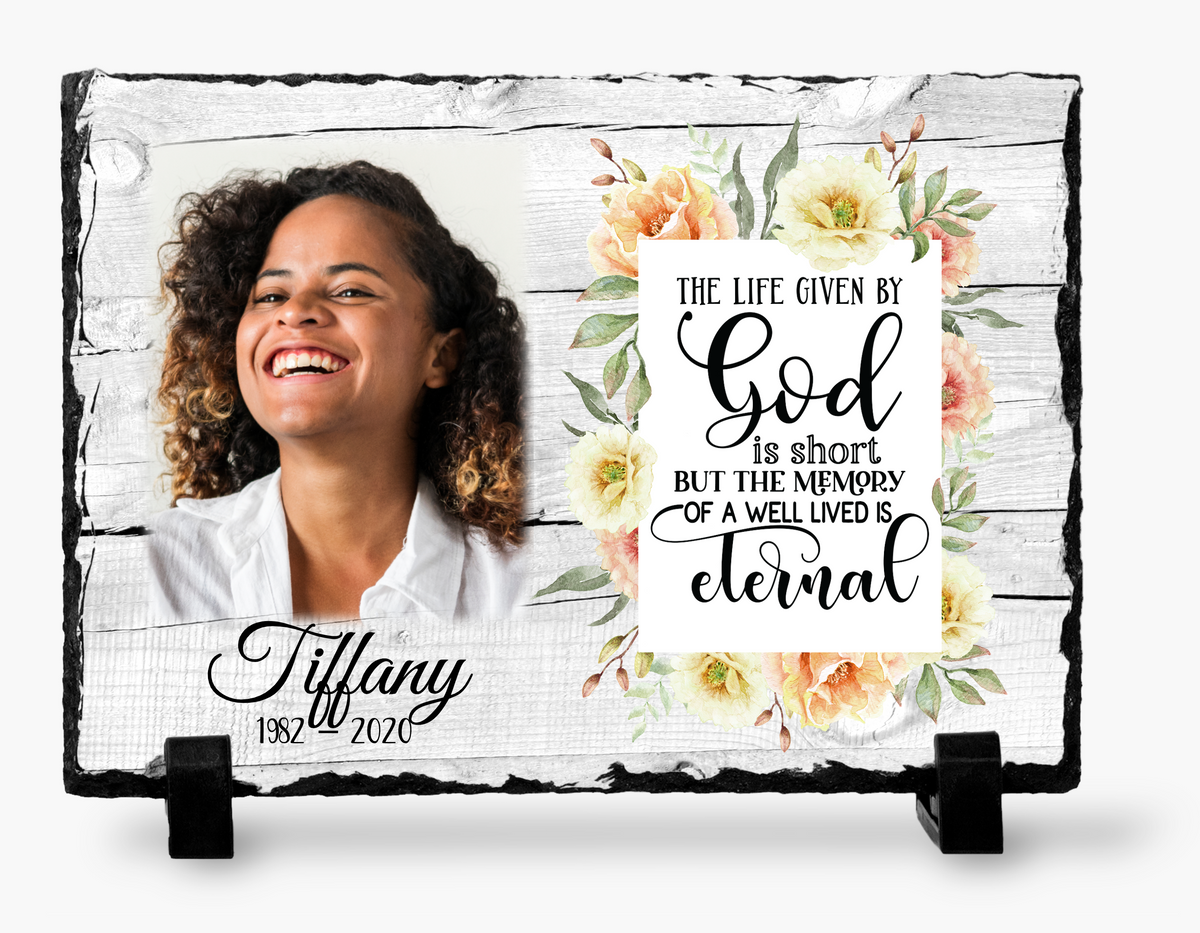 Floral memorial photo slate with a woman’s portrait, religious life-and-memory tribute, sample name and dates, and black stands
